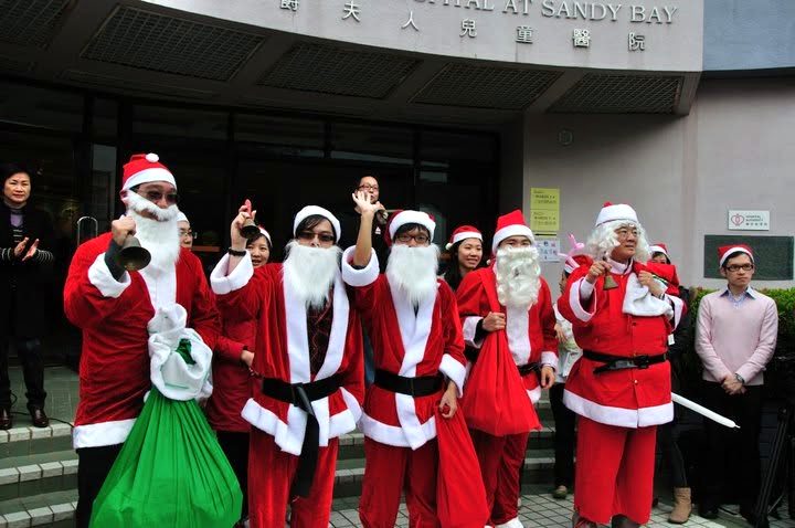 A row of people in Santa Claus suits and white beards stand on the steps outside a building entrance; some wave, one rings a hand bell, and a green sack sits in front of them.