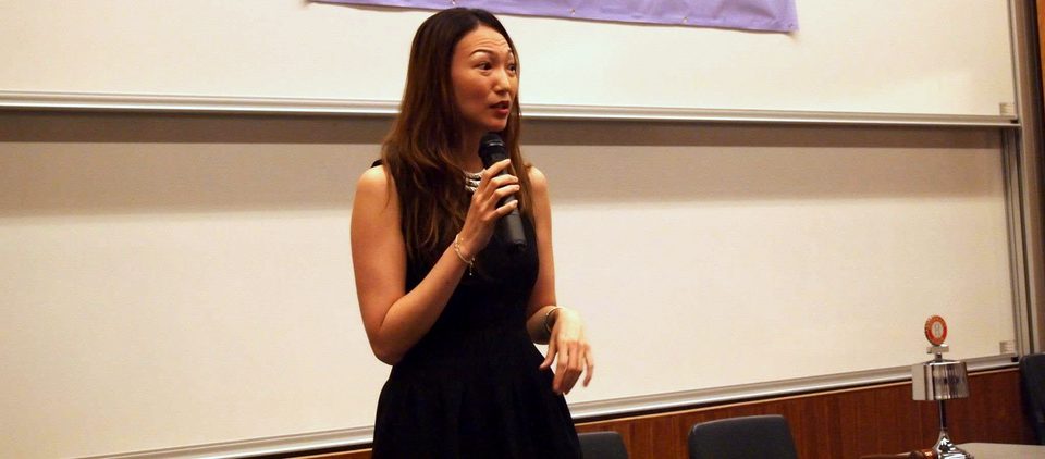 A speaker in a black dress holds a microphone in front of a whiteboard.
