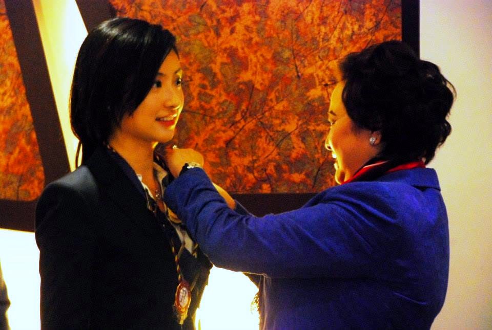 One person in a blue jacket fastens a ceremonial collar round the neck of another, who is smiling.