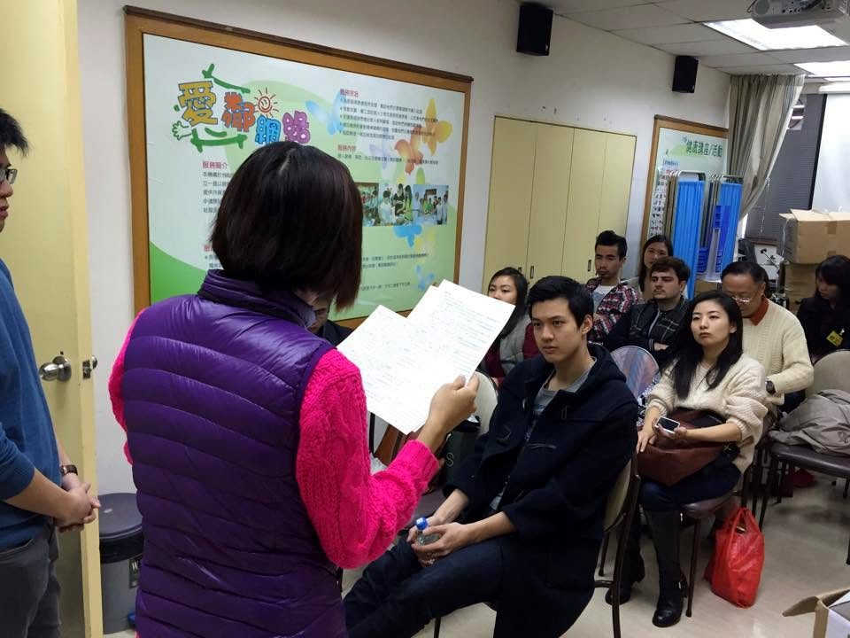 A person in a purple padded waistcoat, seen from behind, reads from sheets of paper to a room of seated people who are listening.
