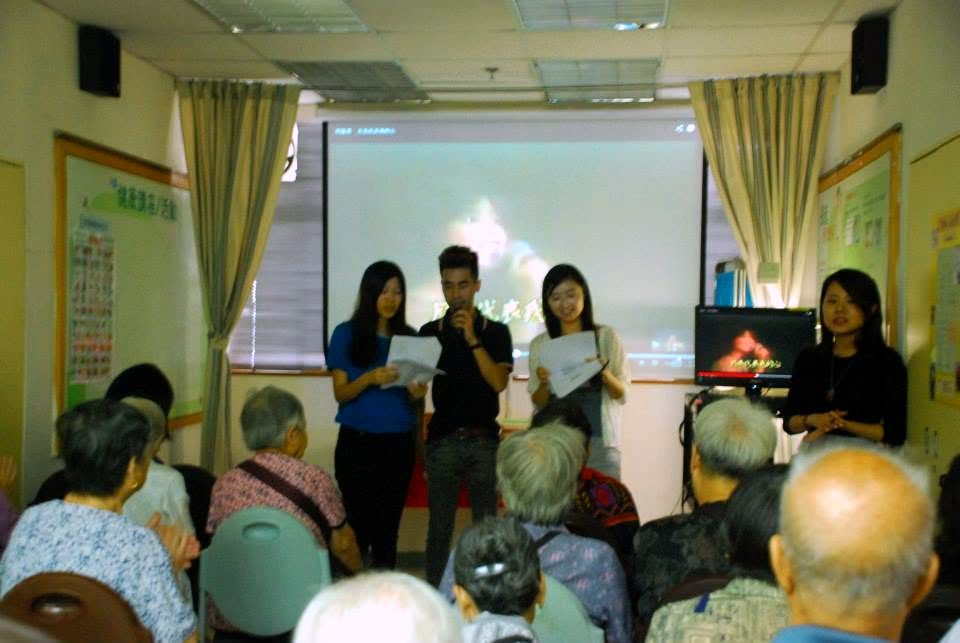 Four people stand at the front of a room, two holding sheets of paper and one a microphone, facing rows of seated people seen from behind.