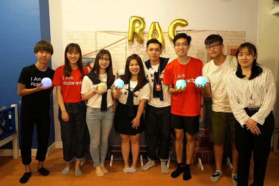 Eight people stand in a row under gold balloons that spell RAC, several holding small glowing globes.