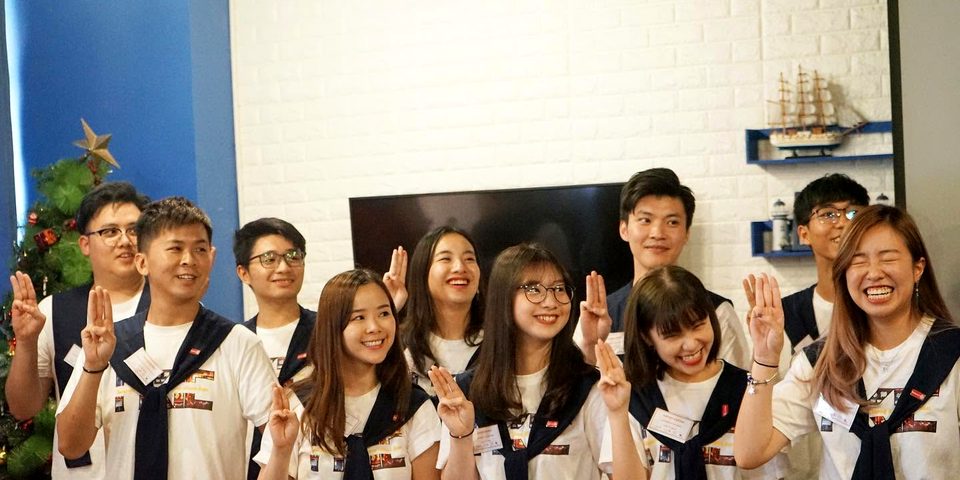 Ten people in white T-shirts with navy scarves stand in two rows, each raising three fingers of one hand; several are laughing.