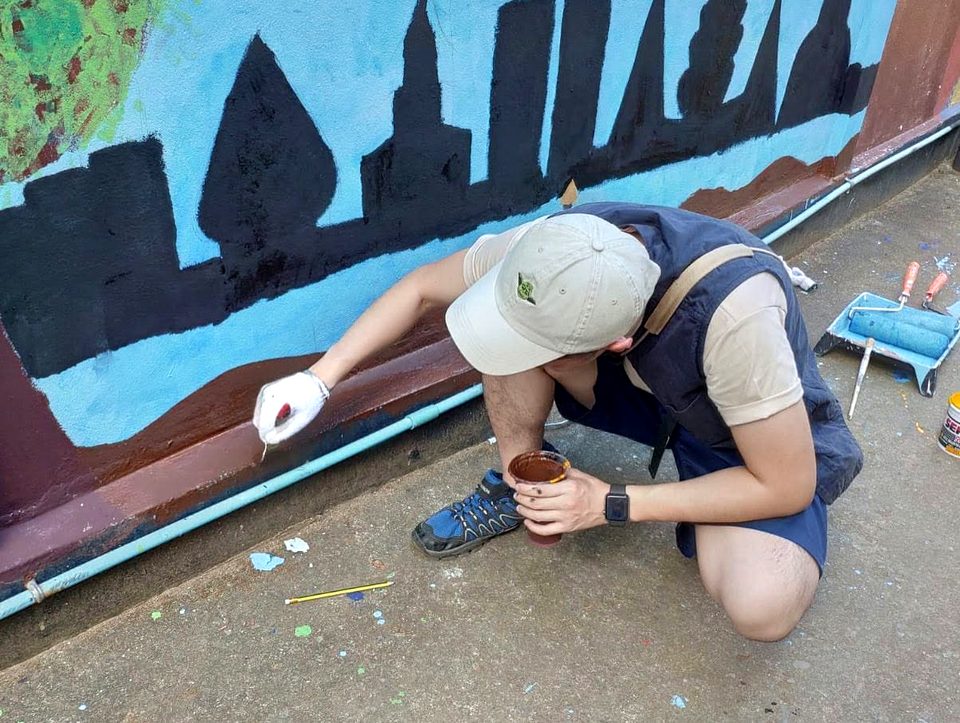 A person in a cap and a work glove crouches to paint the foot of a mural with a fine brush, holding a cup of paint; a roller and tray lie beside them.