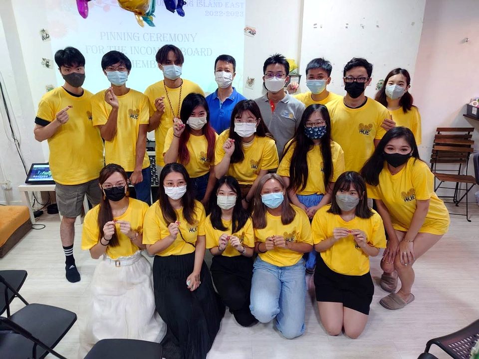 About twenty people in face masks, most in yellow T-shirts, pose in rows in front of a projection screen.
