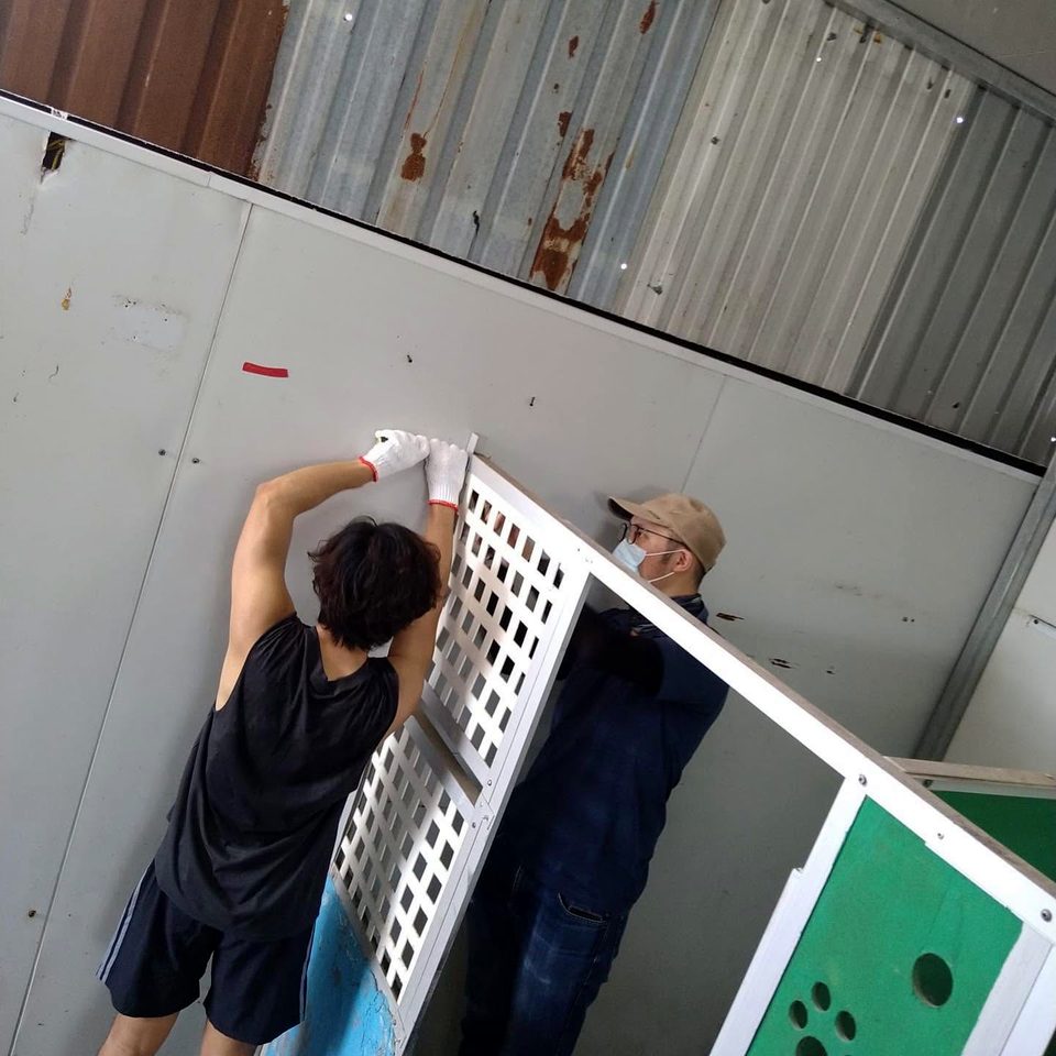 Two people fit a white lattice panel to the front of a kennel; one, seen from behind, wears work gloves, and the other a cap and face mask.